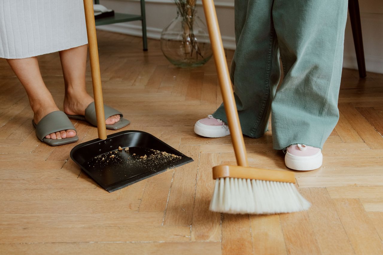 Gros plan sur des personnes nettoyant un sol en bois à l’intérieur avec un balai et une pelle.